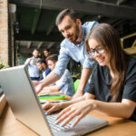 Personas trabajando en un espacio de oficina moderno y colaborativo. En primer plano, una persona utiliza un ordenador portátil sobre una mesa de madera, mientras otra señala algo en la pantalla. Al fondo, se observan más personas reunidas y elementos como paredes de ladrillo, grandes ventanales y mobiliario verde, que transmiten un ambiente abierto y creativo.