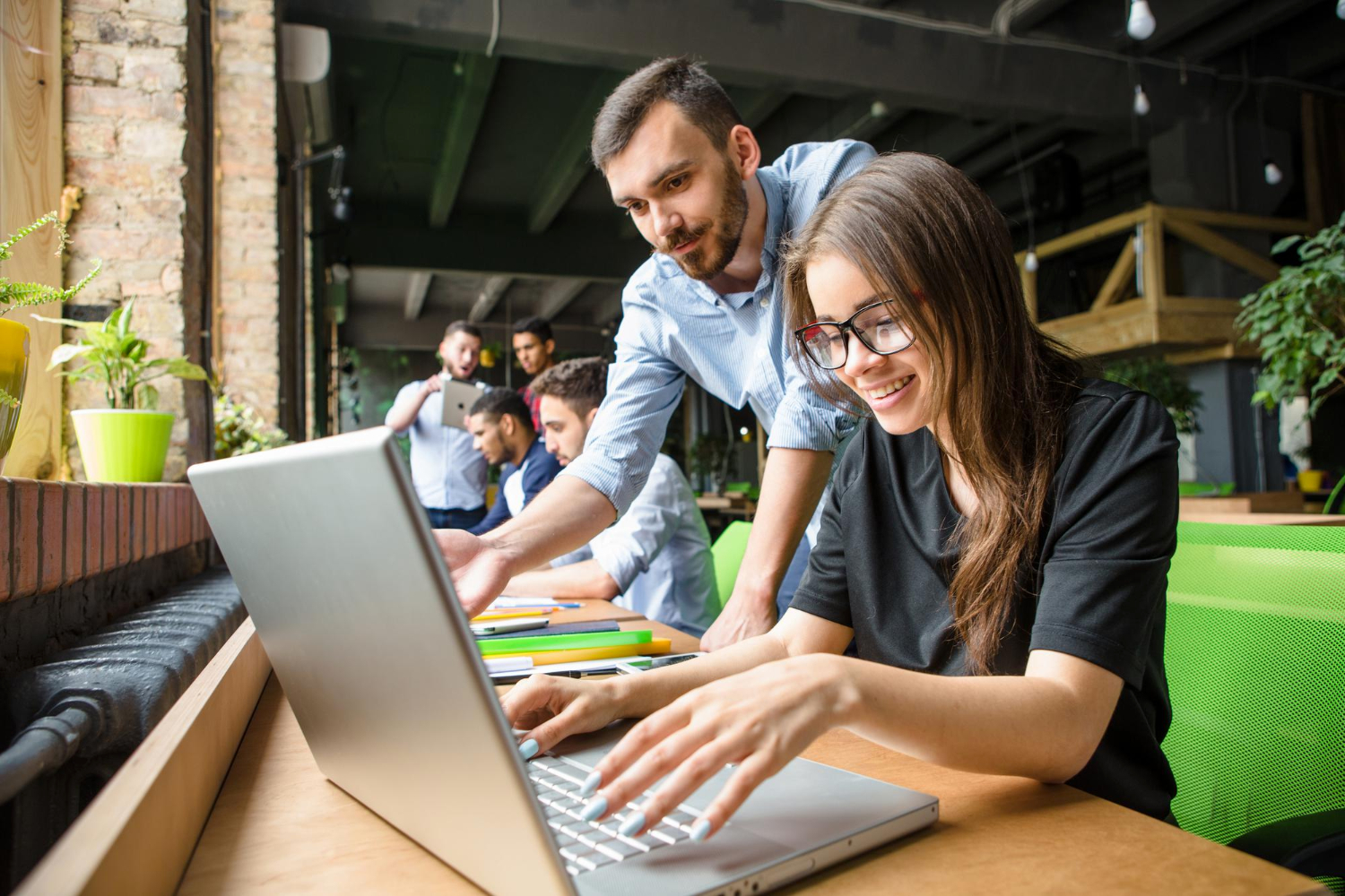 Personas trabajando en un espacio de oficina moderno y colaborativo. En primer plano, una persona utiliza un ordenador portátil sobre una mesa de madera, mientras otra señala algo en la pantalla. Al fondo, se observan más personas reunidas y elementos como paredes de ladrillo, grandes ventanales y mobiliario verde, que transmiten un ambiente abierto y creativo.